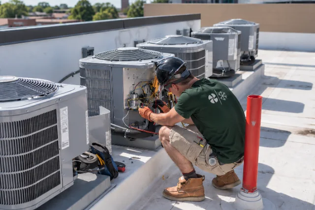 A technician crouched at an open rooftop condensing unit with gauges on the lines, a row of units behind him
